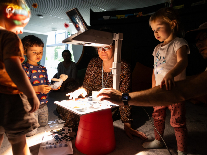 W sali panuje półmrok. Dzieci wraz z edukatorką oglądają przedmioty pod lampą.