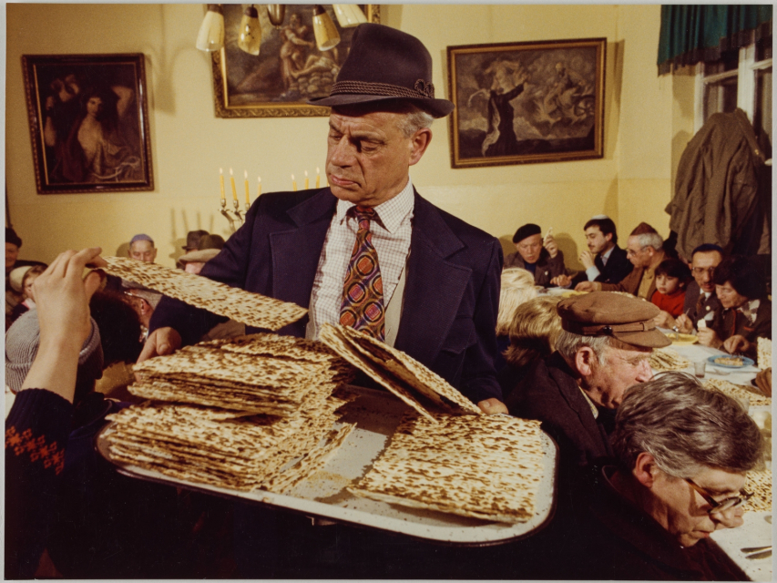 Mężczyzna w kapeluszu trzyma przed sobą talerz z pieczonymi macami. W tle grupa osób siedząca przy stole podczas święta Pesach.