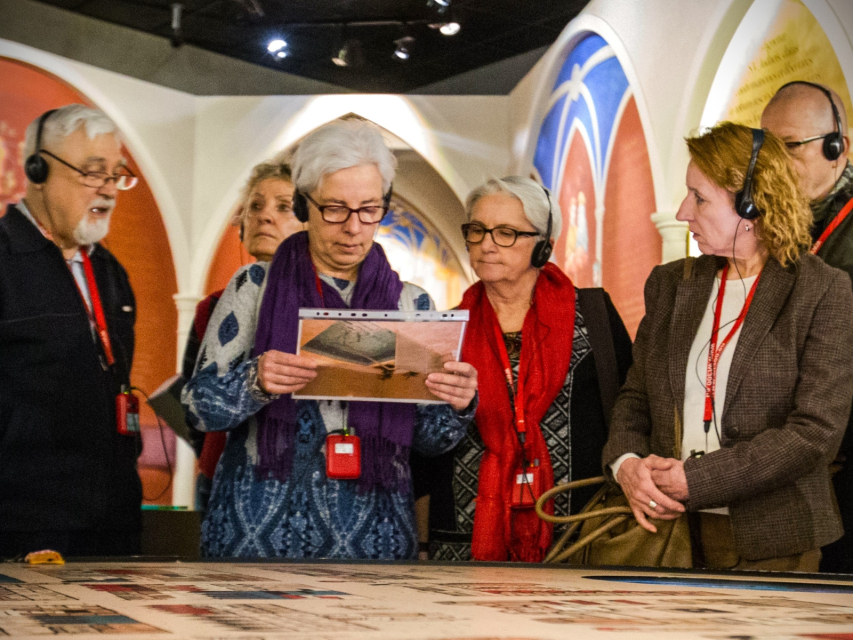 Grupa seniorów stoi w galerii "Pierwsze spotkania" na wystawie stałej w Muzeum POLIN. Kobieta stojąca w środku trzyma jakieś zdjęcie w plastikowej koszulce.