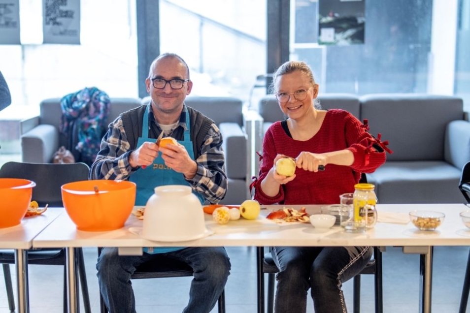 Za stołem kuchennym siedzą dwie osoby, radośnie obierają owoce. Przed nimi na stole pomarańczowe i białe miski oraz pokrojone owoce