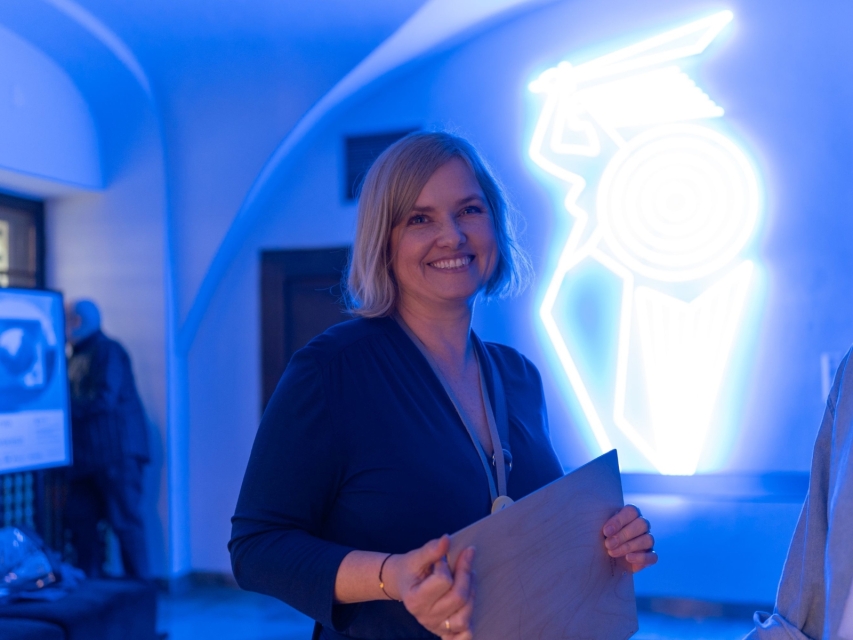 Uśmiechnięta Monika Michałek na tle ściany z neonową warszawską syrenką.