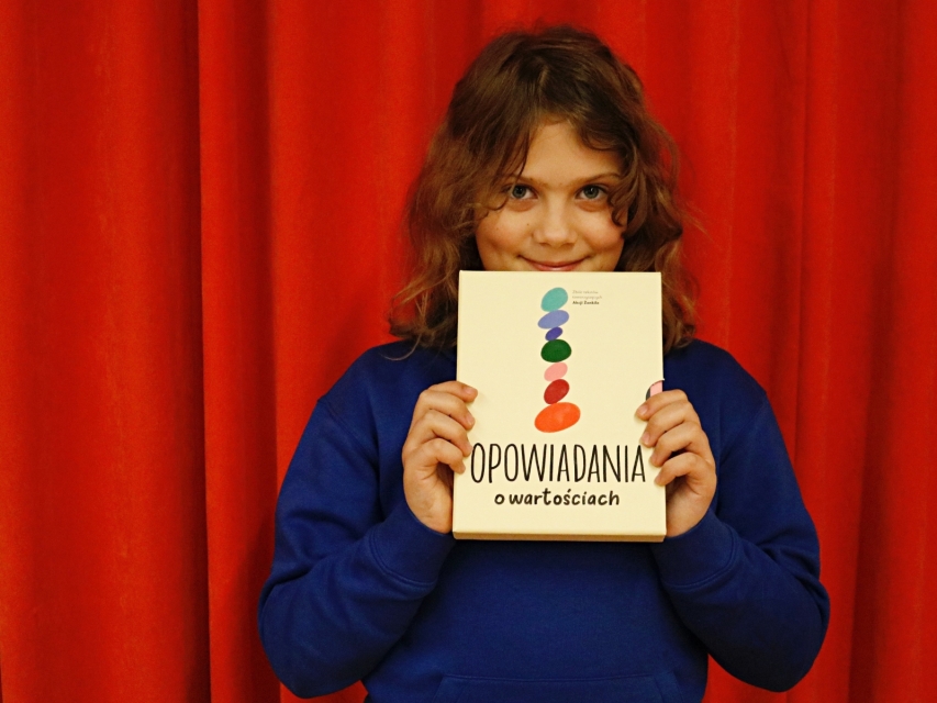 Uśmiechnięta dziewczynka trzyma przed sobą egzemplarz "Opowiadań o wartościach".