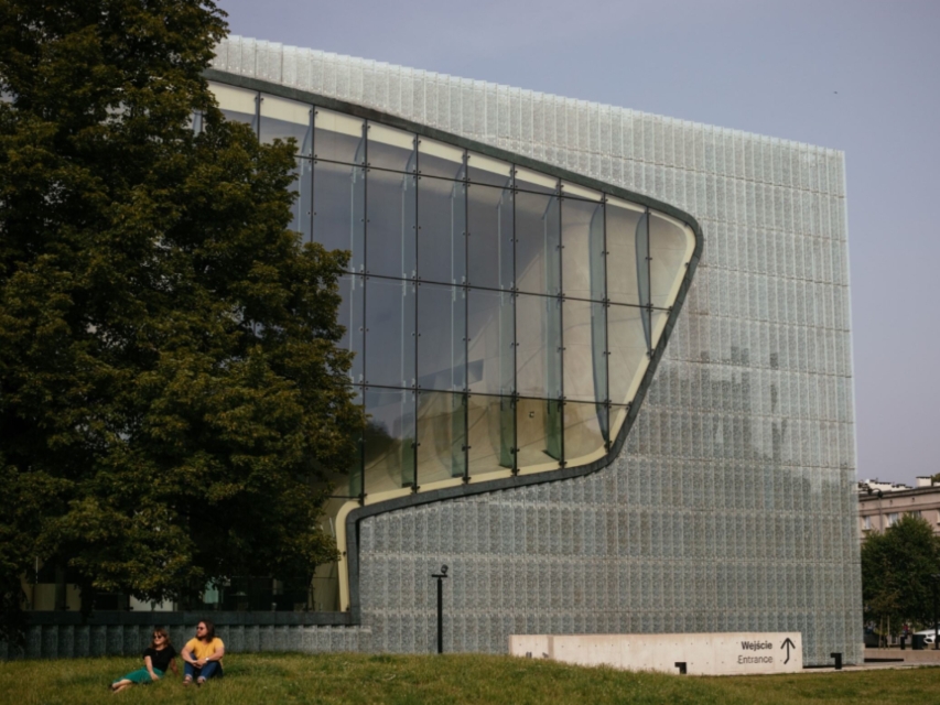 Kobieta i mężczyzna siedzą na łące przy budynku Muzeum POLIN.