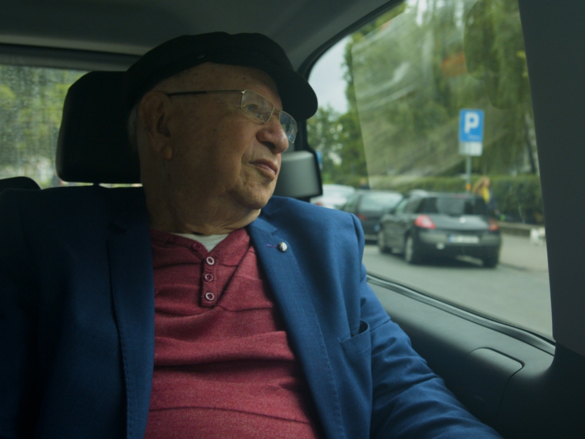 Kadr z filmu "Wśród sąsiadów". Starszy mężczyzna jedzie samochodem jako pasażer.