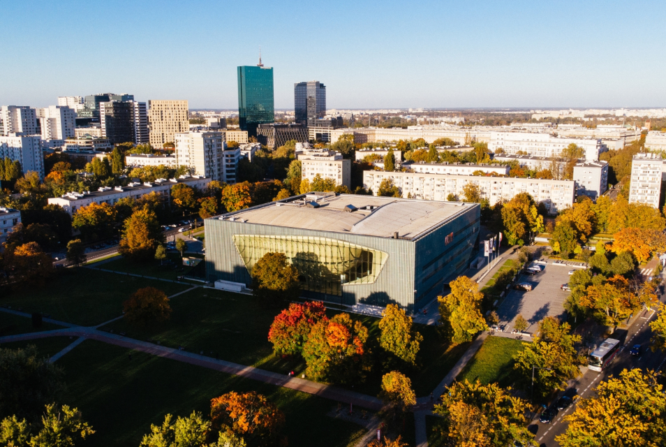 Budynek Muzeum POLIN pokazany z góry, stoi wśród drzew, bloków i wieżowców oświetlony przez słońce.