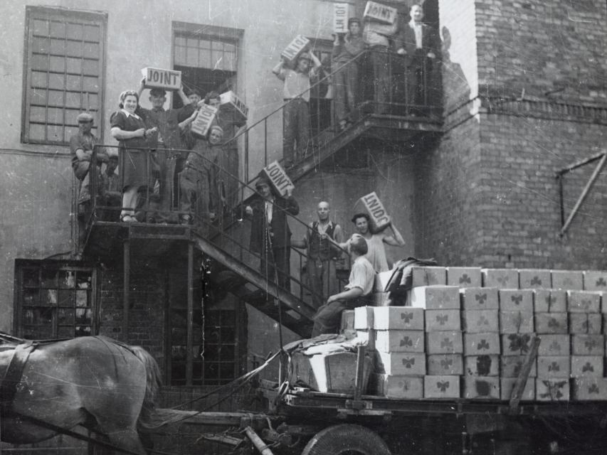 Supplies being taken off a horse-drawn wagon and being carried into a warehouse in Warsaw; the crates are labeled "JOINT".