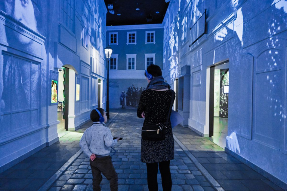 Kobieta z dzieckiem w słuchawkach spacerują po galerii "Na żydowskiej ulicy" na wystawie stałej w Muzeum POLIN.