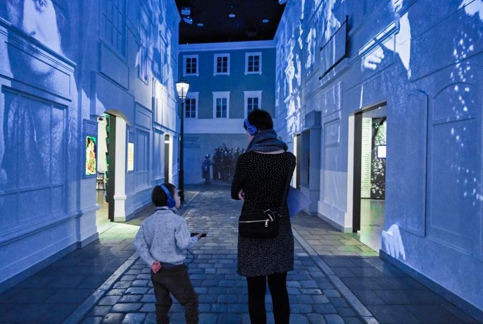 A woman with a child wearing headphones strolls through the "On a Jewish Street" gallery at the Core Exhibition at POLIN Museum.