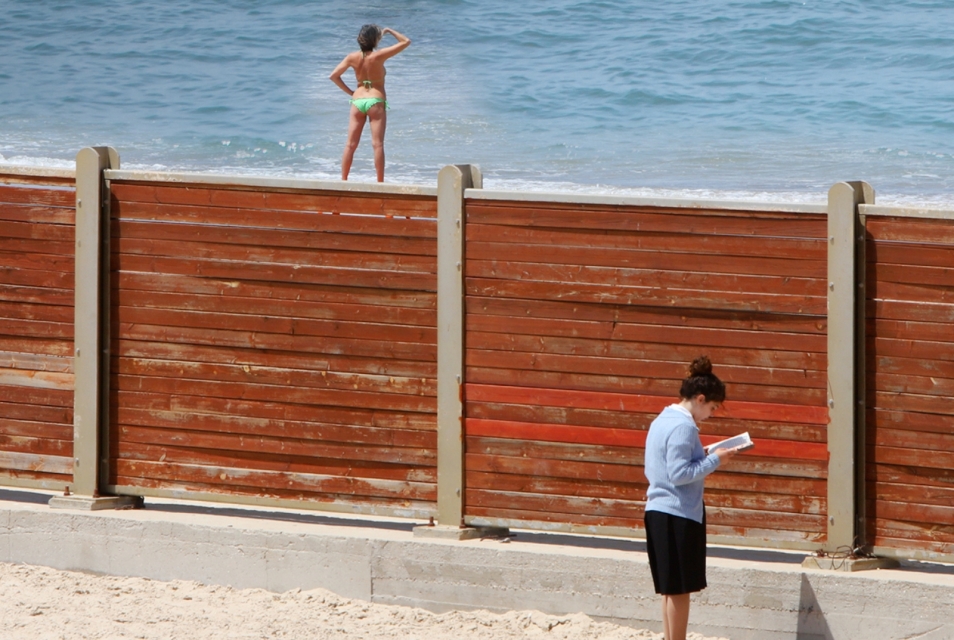 Kadr z fimu "Koszerna plaża". Kobieta ubrana w sweter i spódnicę czyta książkę na plaży. Za murem stoi inna kobieta, ubrana tylko w strój kąpielowy.