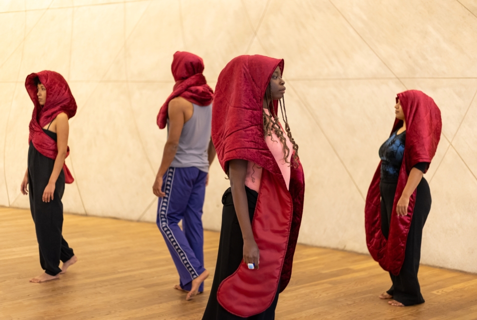 Three dark-skinned women and one dark-skinned man. They all wear red hanging headdresses.