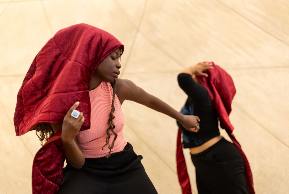 Two dark-skinned women dancing. Both wear red hanging headdresses.