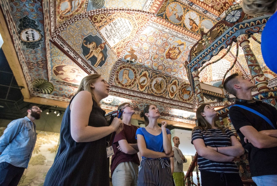 Grupa młodych osób stoi z przewodnikiem na wystawie stałej w Muzeum POLIN, przy rekonstrukcji synagogi z Gwoźdźca.