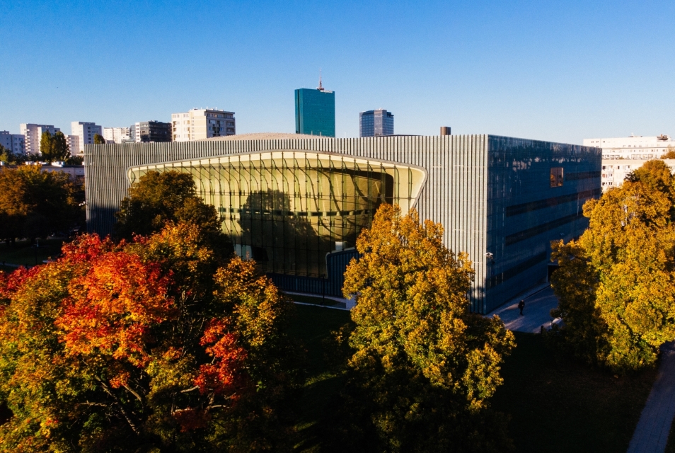 The POLIN Museum building surrounded by autumn trees. Skyscrapers in the background.