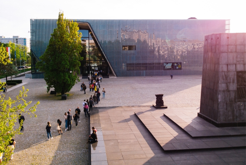 Kolejka osób czeka na wejście do budynku Muzeum POLIN. Świeci słońce. Z prawej strony widoczny pomnik Bohaterów Getta.