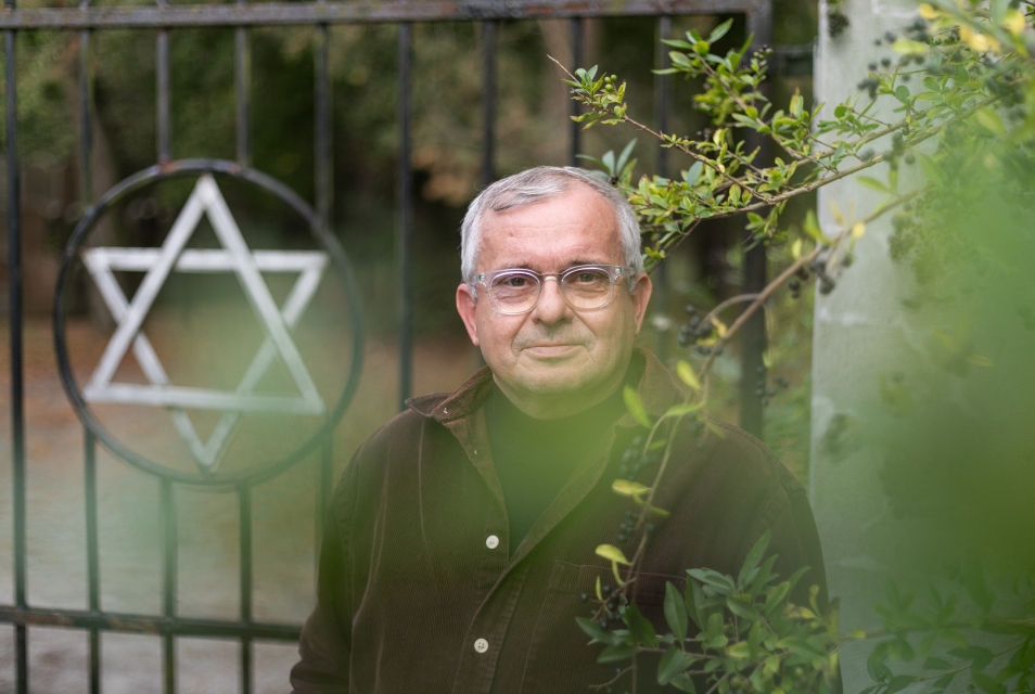 Andrzej Białek widziany zza zielonych krzewów. Za nim ogrodzenie żydowskiego cmentarza z gwiazdą Dawida