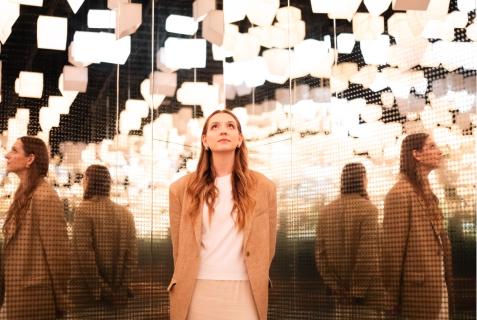 A woman with her head raised stands in the Bonds Gallery under a ceiling made of hanging lamps shaped like houses.