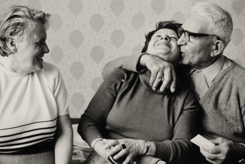 The woman on the left, Pola Najder-Gałkiewicz (in the middle) and Andrzej Śliwiak (on the right) are sitting next to one another inside a flat. The man has his arm firmly around the woman sitting in the middle, kissing her on the cheek; the other woman is watching and smiling. Patterned wallpaper can be seen on the wall in the background.