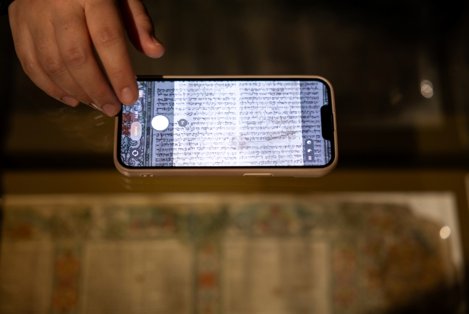 Ktoś fotografuje telefonem pergamin zapisany w języku hebrajskim. Przykłada telefon do szyby gabloty.