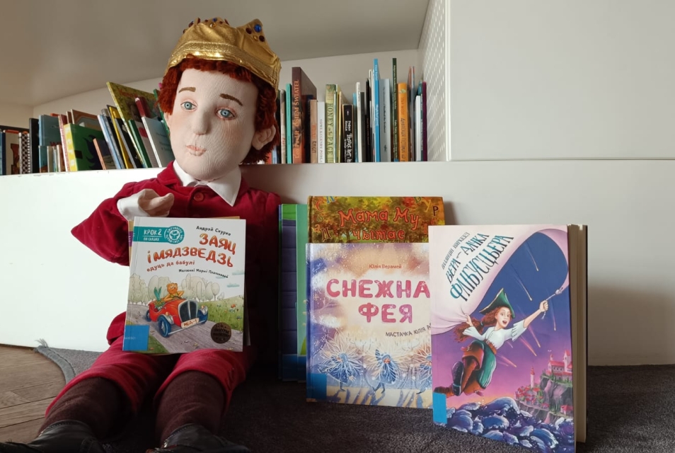 Лялька Кароль Мацюсь сядзіць каля палічкі з кнігамі ў атачэнні беларускіх кніг