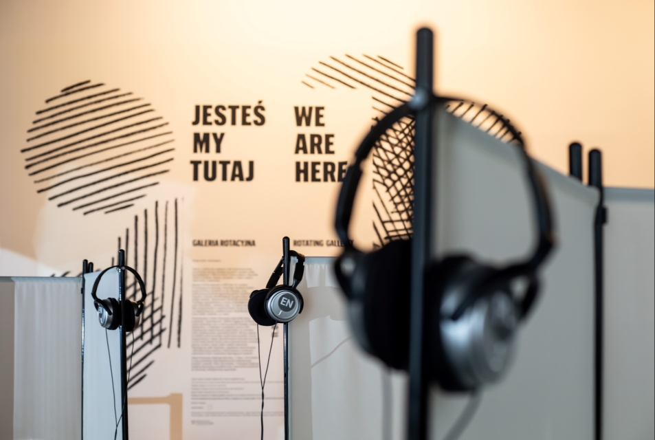 Słuchawki wiszą na kołkach w Galerii Rotacyjnej. Na ścianie napisy: jesteśmy tutaj, we are here.