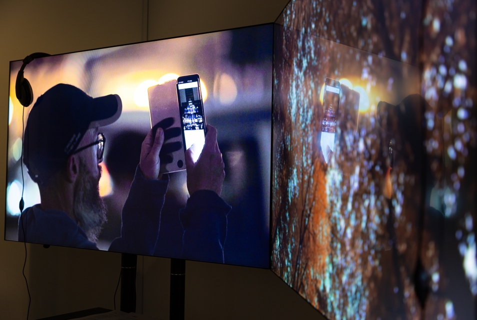 The photo shows two LED screens. On one of them, a man is taking a photo with his phone, and on the other, tree branches are visible. A pair of headphones is hanging on one of the screens.