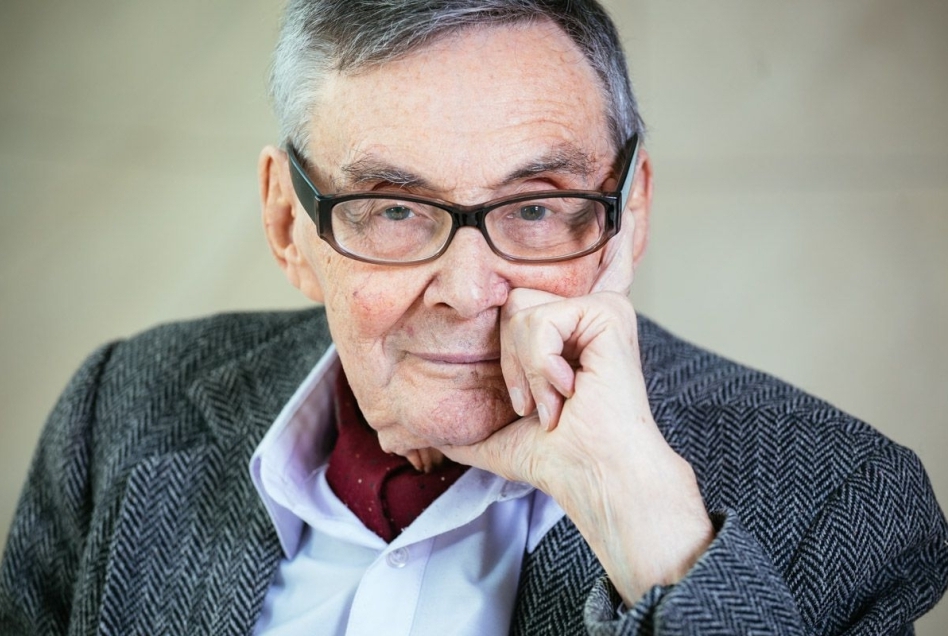 Marian Turski ubrany w niebieską koszulę i marynarkę. Podpiera twarz dłonią i patrzy przed siebie.