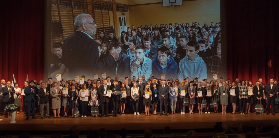 Duża grupa ludzi stoi na scenie. W tle wyświetla się zdjęcie mężczyzny i grupy młodych osób.
