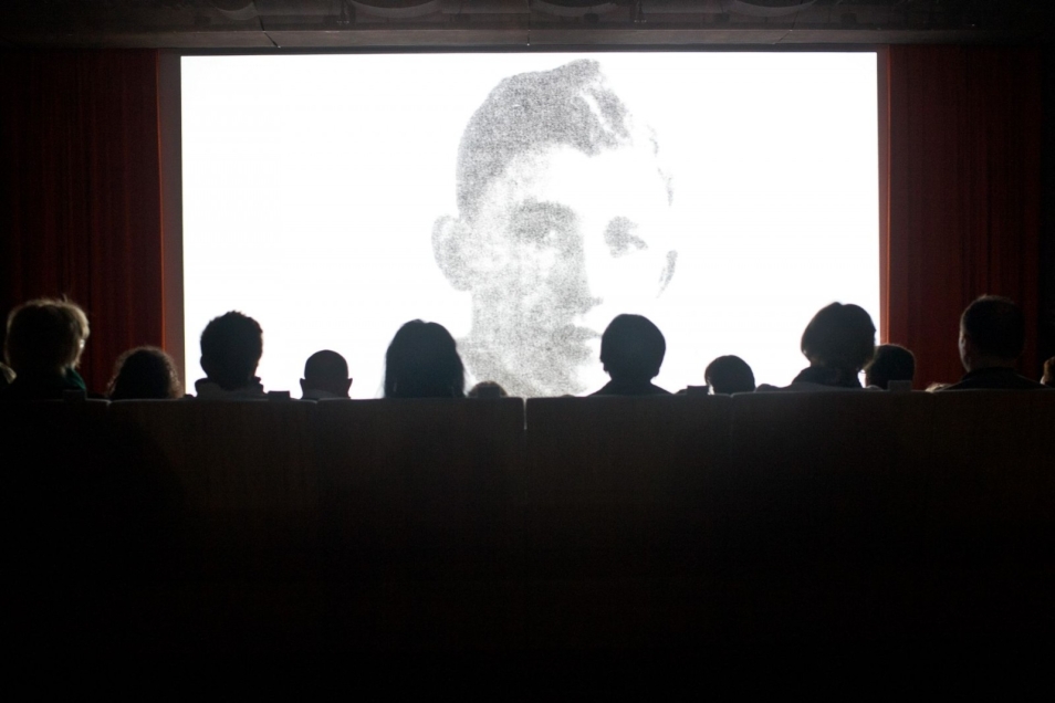 Grupa ludzi ogląda czarnobiały film. Kadr filmu przedstawia młodego mężczyznę patrzącego na wprost.
