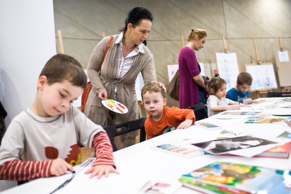 Warsztaty plastyczne wewnątrz budynku muzeum podczas "Dnia Dziecka" 2013. Dwie kobiety pomagają dzieciom przy stole z pracami i materiałami plastycznymi.