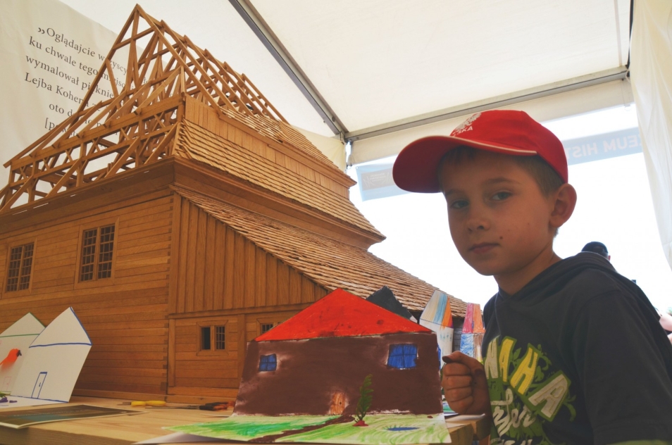 Chłopiec ze swoją pracą na tle drewnianego modelu synagogi.