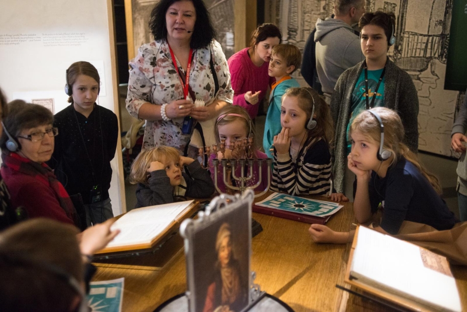 SZKOŁA PODSTAWOWA 4-6: Między synagogą a domem rodzinnym. święta i uroczystości żydowskie. Na zdjęciu: w środku przewodniczka stoi przed stołem, na którym stoja przedmioty związane z codziennym życiem religijnej żydowskiej rodziny. Wokół stołu stoją dzieci i słuchają opowieści przewodniczki.