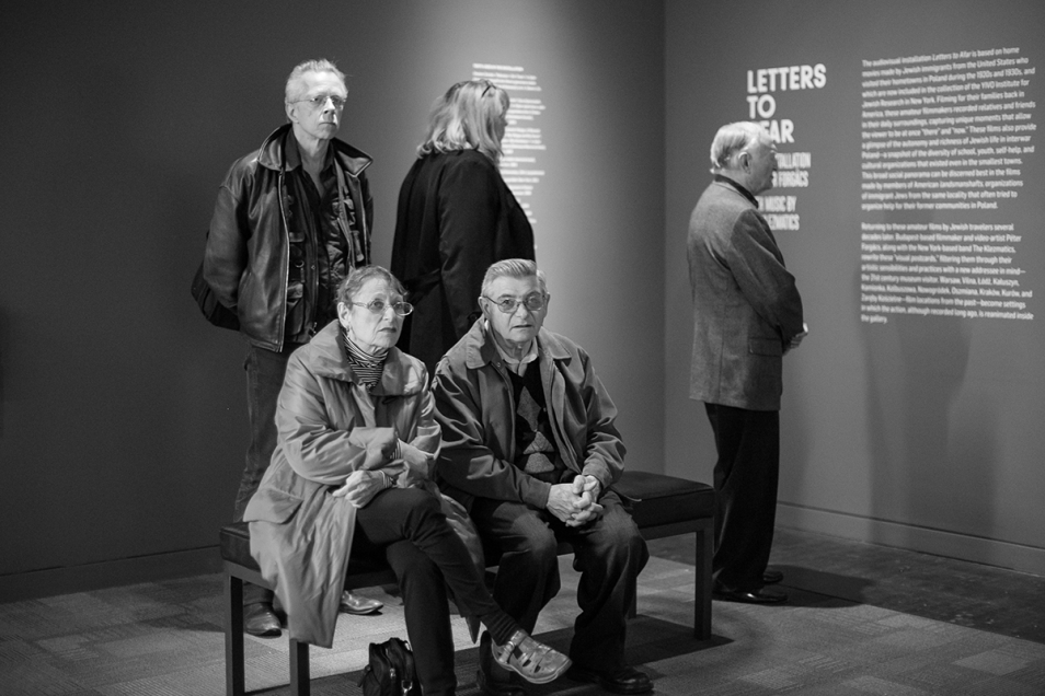 Kilka osób przy ekspozycji "Letters to Afar". Dwie osoby siedzą na ławce, trzy stoją przy ścianach z opisami.
