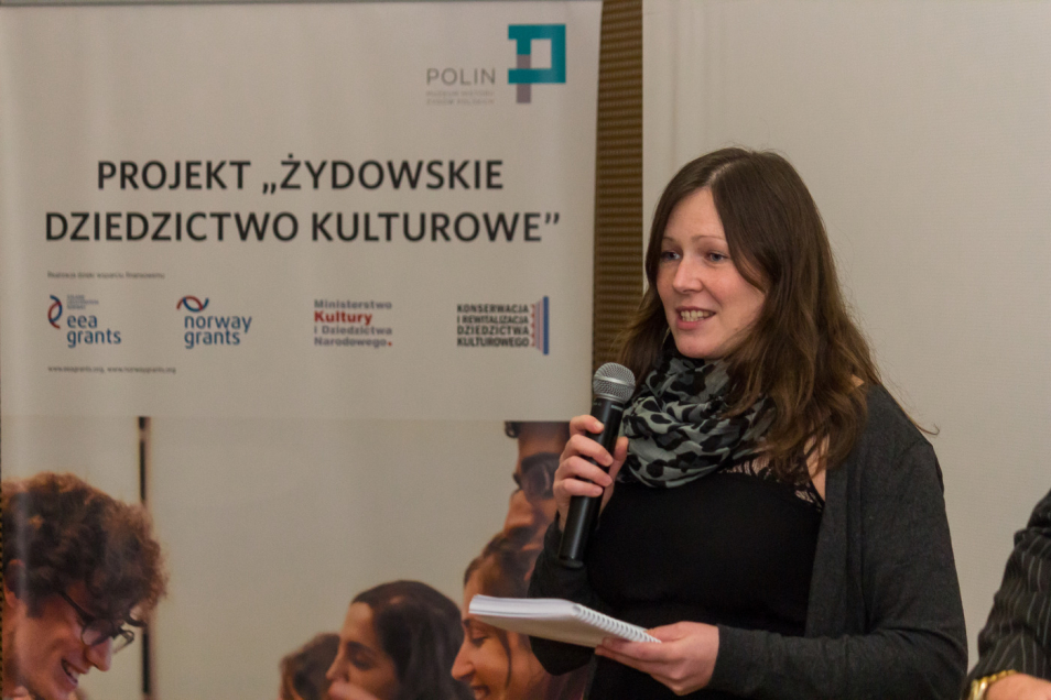Kobieta mówi przez mikrofon. W tle baner z napisem projekt Żydowskie Dziedzictwo Kulturowe.