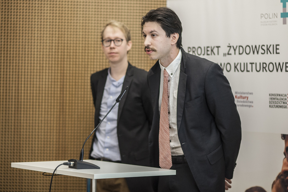Dwóch mężczyzn przy mównicy. W tle plansza z napisem projekt "Żydowskie Dziedzictwo Kulturowe".