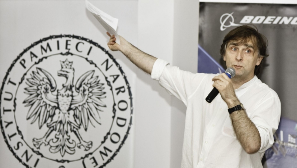 Mężczyzna mówi przez mikrofon i pokazuje logo Instytutu Pamięci Narodowej.