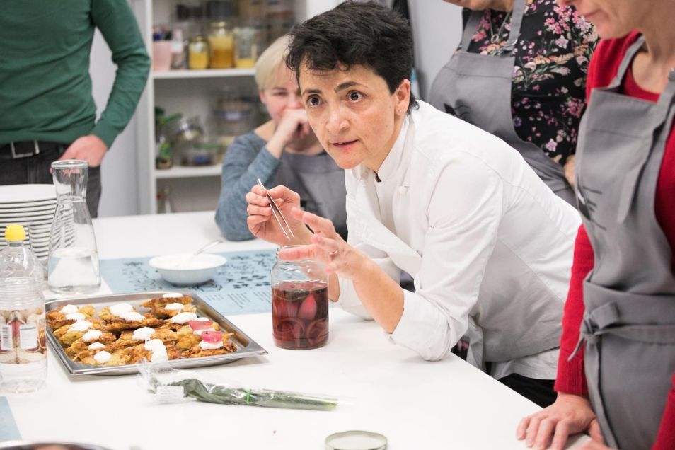 Alessia di Donato w białym fartuchu pochyla się nad stołem i tłumaczy coś uczestnikom i uczestniczkom warsztatów.