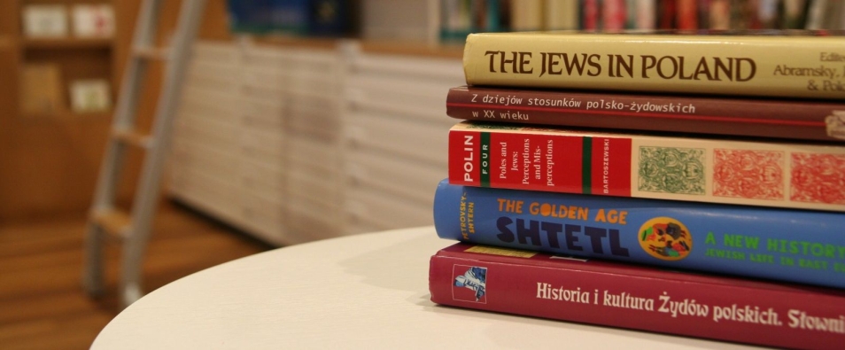 Grzbiety książek ułożonych jedna na drugiej. Leżą na blacie stołu. W głębi regały biblioteczne