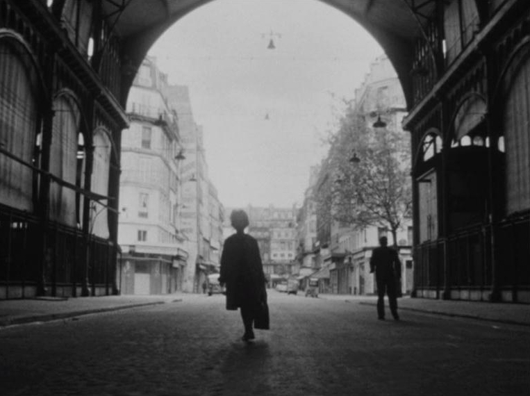 Czarno-biała fotografia. Jakaś postać przechodzi przez tunel. W tle miasto.