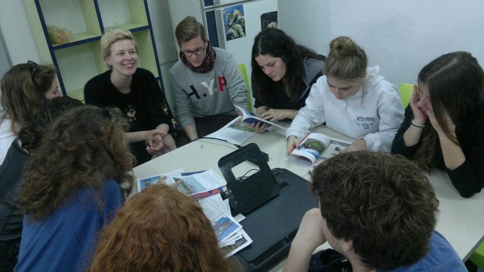 Grupa osób siedzi przy stole podczas warsztatów. Niektórzy przeglądają jakieś fotografie.