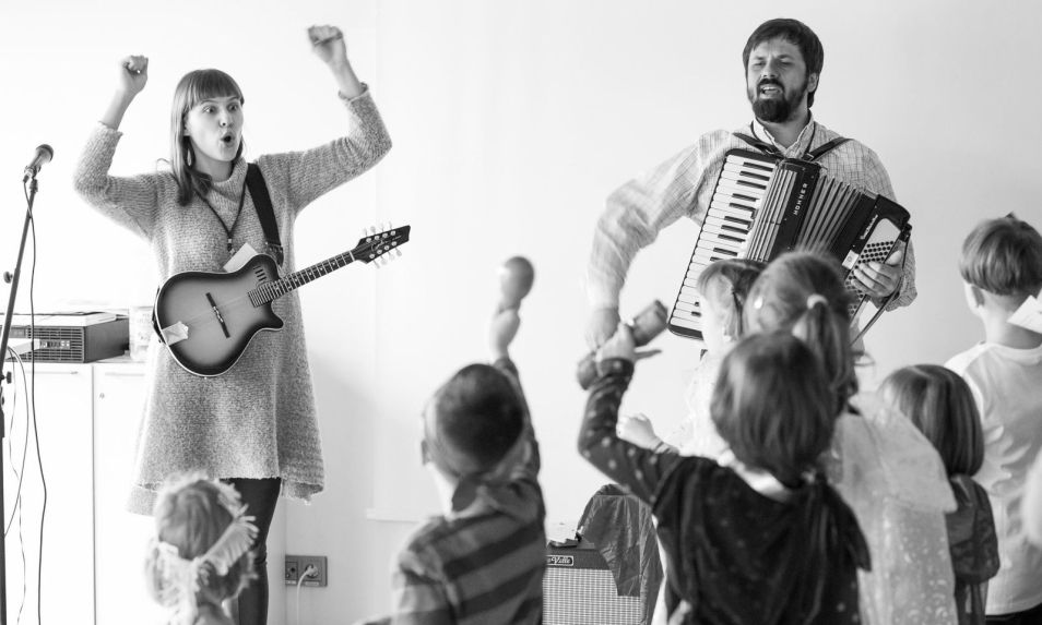 Kobieta i mężczyzna grają na instrumentach. Ich występu słuchają dzieci.