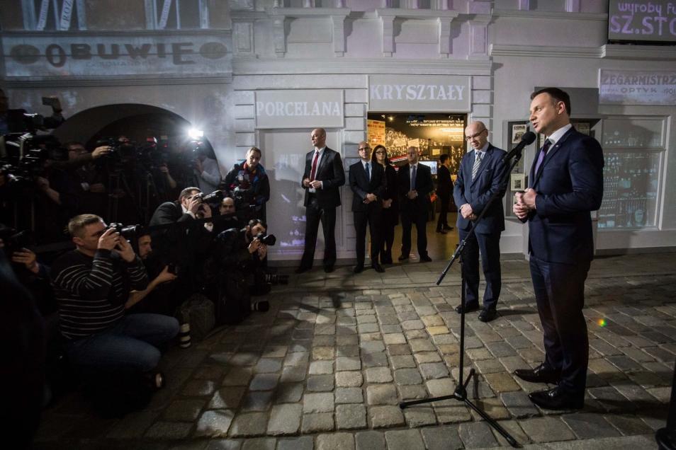  Wystawa stała w muzeum Polin. Część poświęcona 20-leciu międzywojennemu. Przy mikrofonie stoi prezydent Andrzej Duda. Przemawia. Obok stoi dyrektor muzeum, prof. Dariusz Stola. Fotografowie robią zdjęcia. 