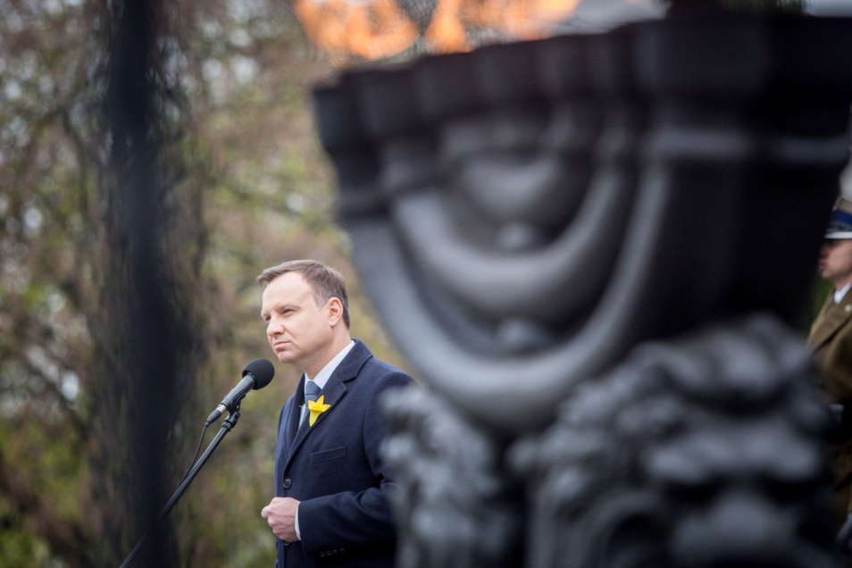 Prezydent Andrzej Duda z żonkilem przypiętym do klapy marynarki przemawia przed mikrofonem na tle Pomnika Bohaterów Getta w Warszawie