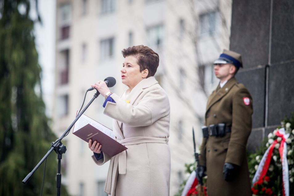 Kobieta stoi przed mikrofonem i czyta przemówienie, obok stoi żołnierz przy wieńcu na którym jest wstęgą z flagą polską