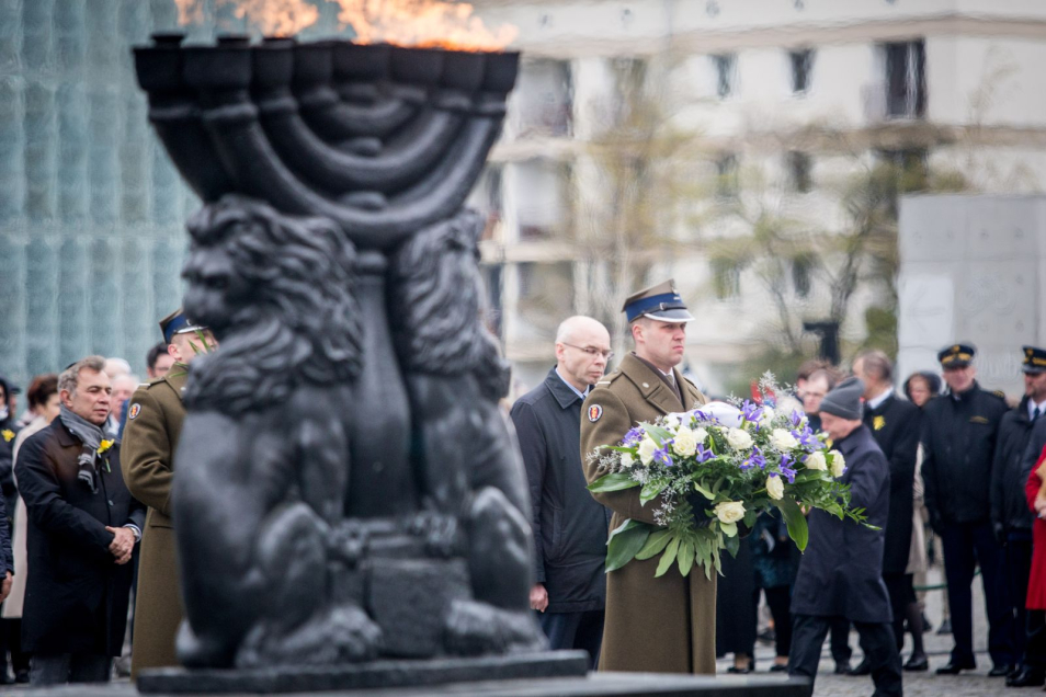 Grupa mężczyzn przed Pomnikiem Bohaterów Getta w Warszawie składa wieńce. Zapalone pochodnie.