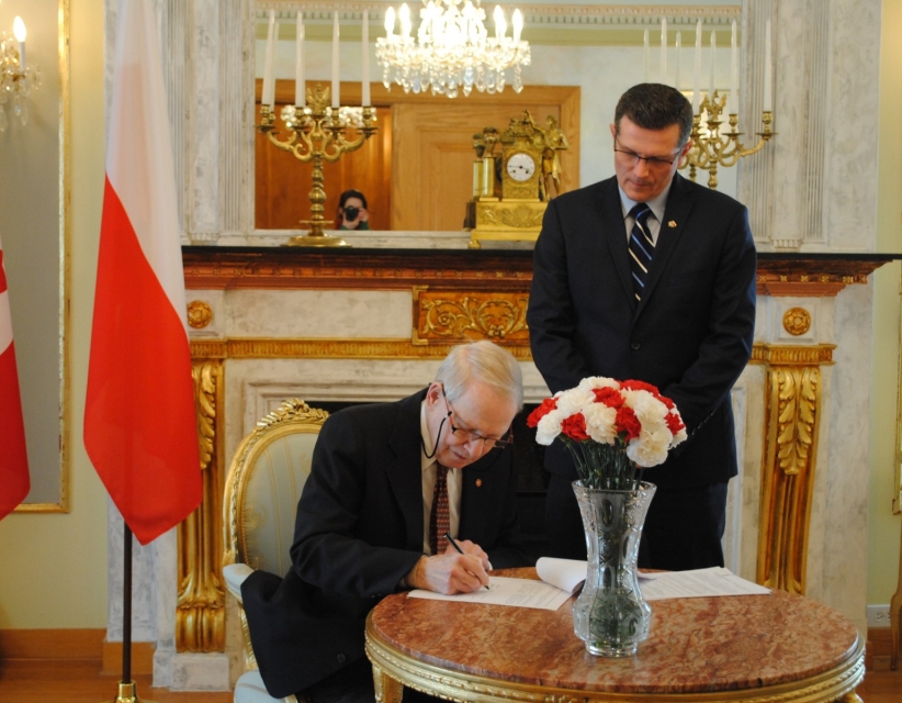 Starszy mężczyzna podpisuje jakiś dokument w gabinecie. Jest tam flaga Polski i biało-czerwony bukiet.