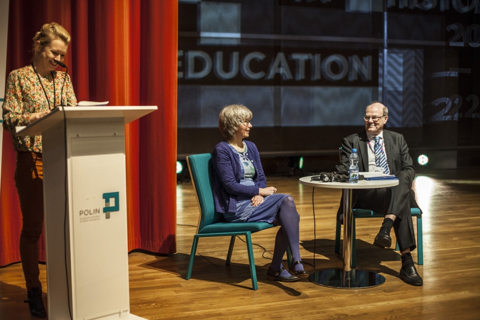 Paneliści siedzą przy stoliku podczas konferencji, jedna kobieta stoi przy mównicy.