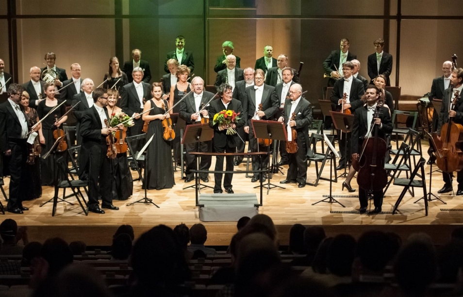 Zakończony koncert. Muzycy orkiestry stoją. Dyrygent w ręku trzyma kwiaty. Stoi przodem do widowni.