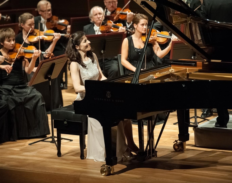 Pianistka w białej sukience gra na fortepianie. Po prawej stronie skrzypaczka w czarnej sukni gra na skrzypcach. 