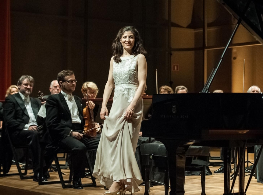Na środku sceny stoi pianistka w białej sukience. Koncert z orkiestrą się zakończył.