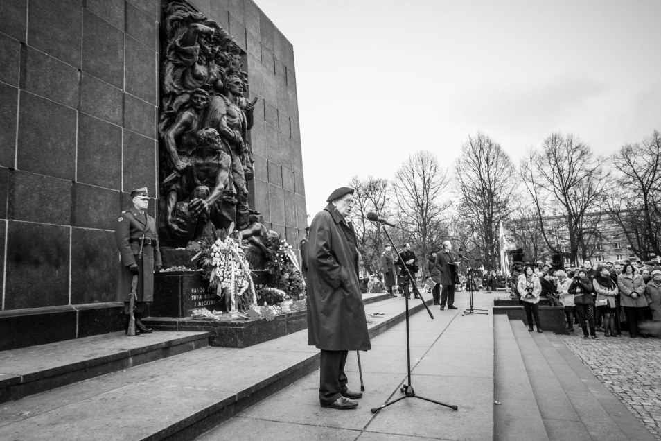 Władysław Bartoszewski przy mównicy. Starszy, łysy mężczyzna w okularach i berecie. Ubrany jest w garnitur.